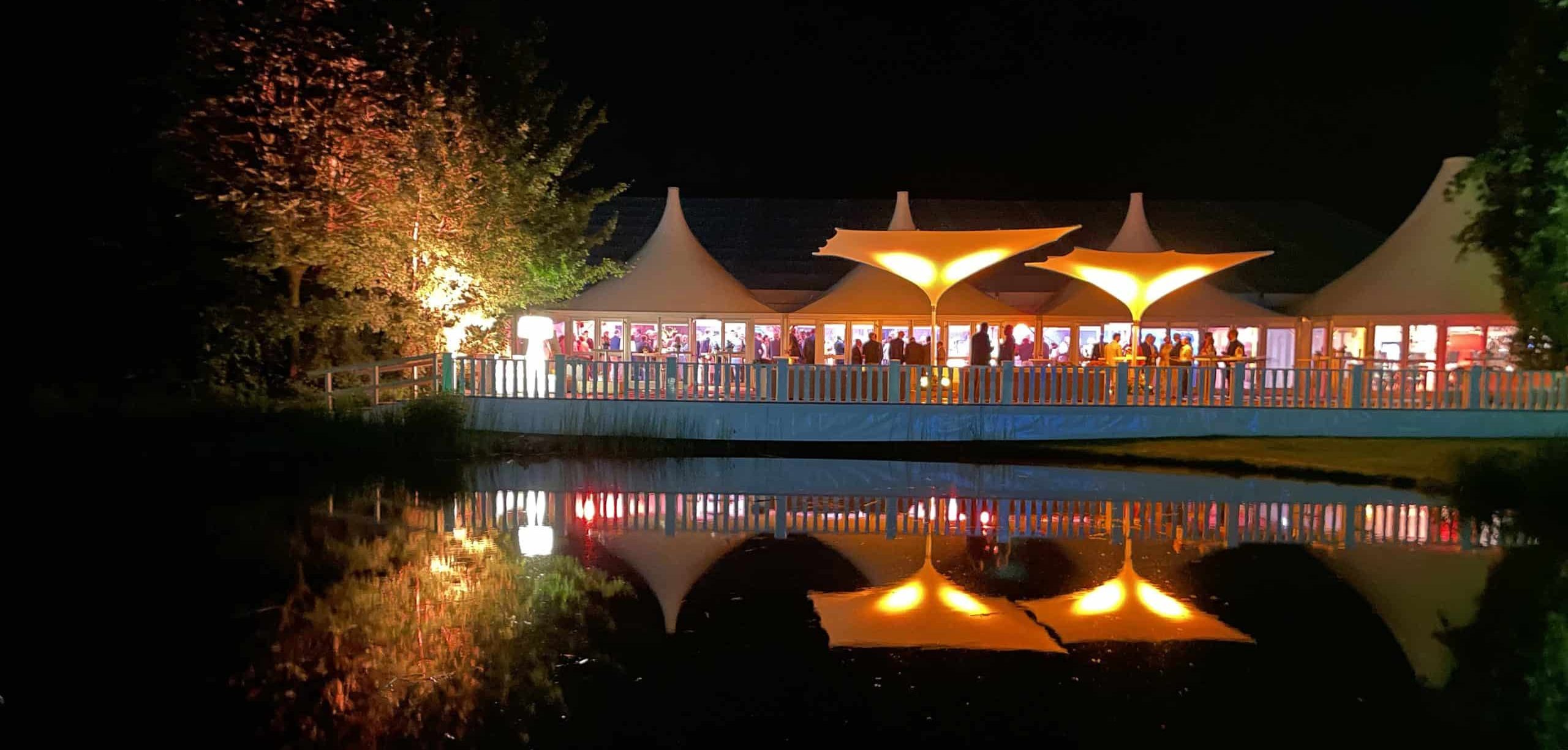 Eventsetting in einer Gartenlandschaft auf einer Terrasse am See, im Hintergrund sind Pagoden und futuristische große Sonnenschirme zu sehen. Gäste unterhalten sich